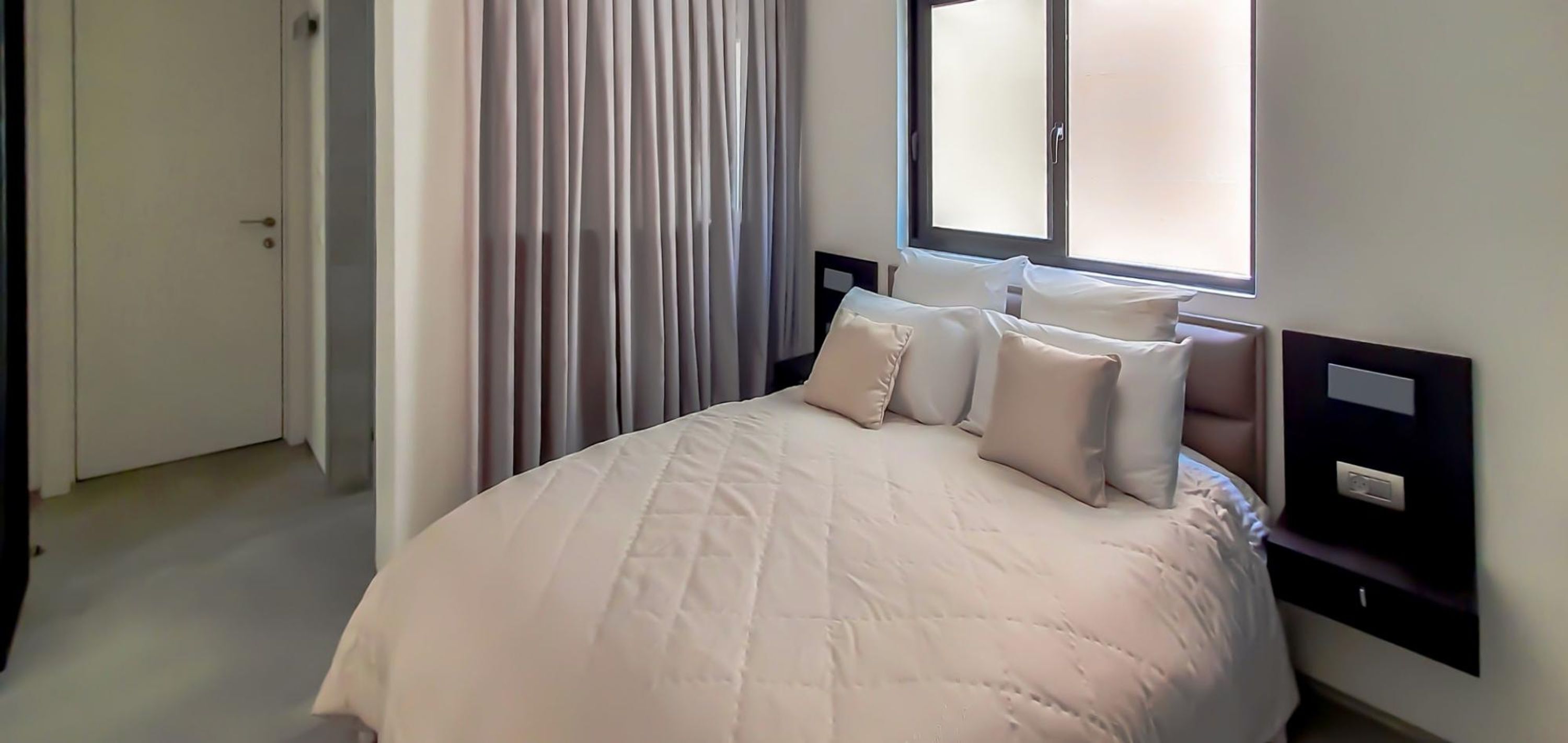 A neatly made bed with a white duvet and pillows, a bedside table with a lamp, and a window with blinds.