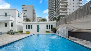 A swimming pool with a waterfall feature, surrounded by white buildings with balconies and a fence. - Puerto Rico Beachside Rental w Pool Otium Rentals (San Juan)
