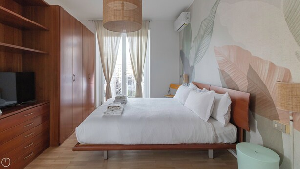 A bedroom with a bed, a television on a wooden cabinet, a window with curtains, and a wall with a leaf pattern. - Italianway - Brianza 14 A (Milan)