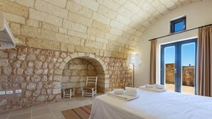 A hotel room with a stone wall, a bed with white linens, a chair, and a view through a glass door. - 2081 Villa Crocilupo by Perle di Puglia (Gagliano del Capo)