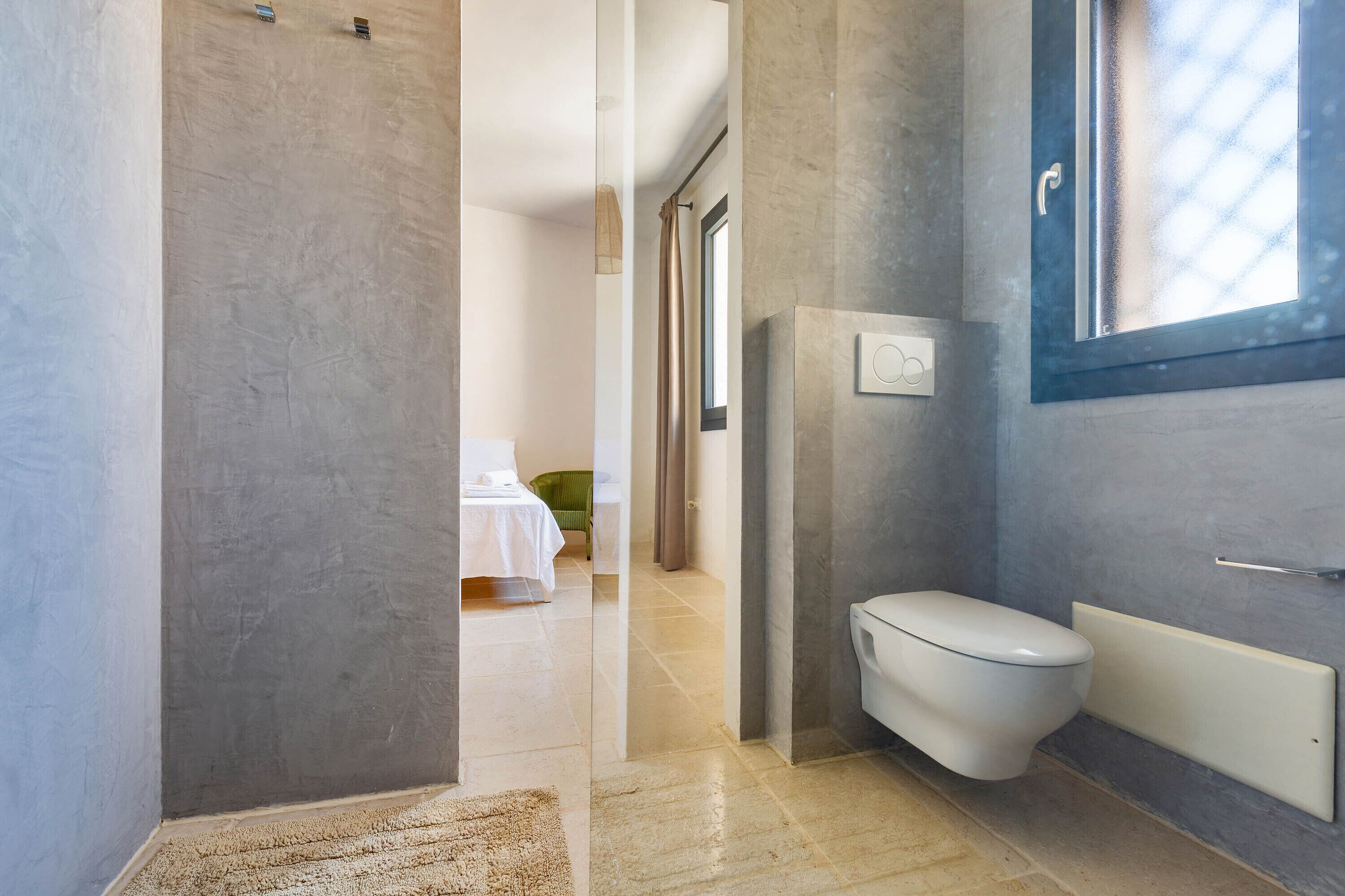 A modern bathroom with a toilet, a shower area, and a visible bed in the adjacent room.