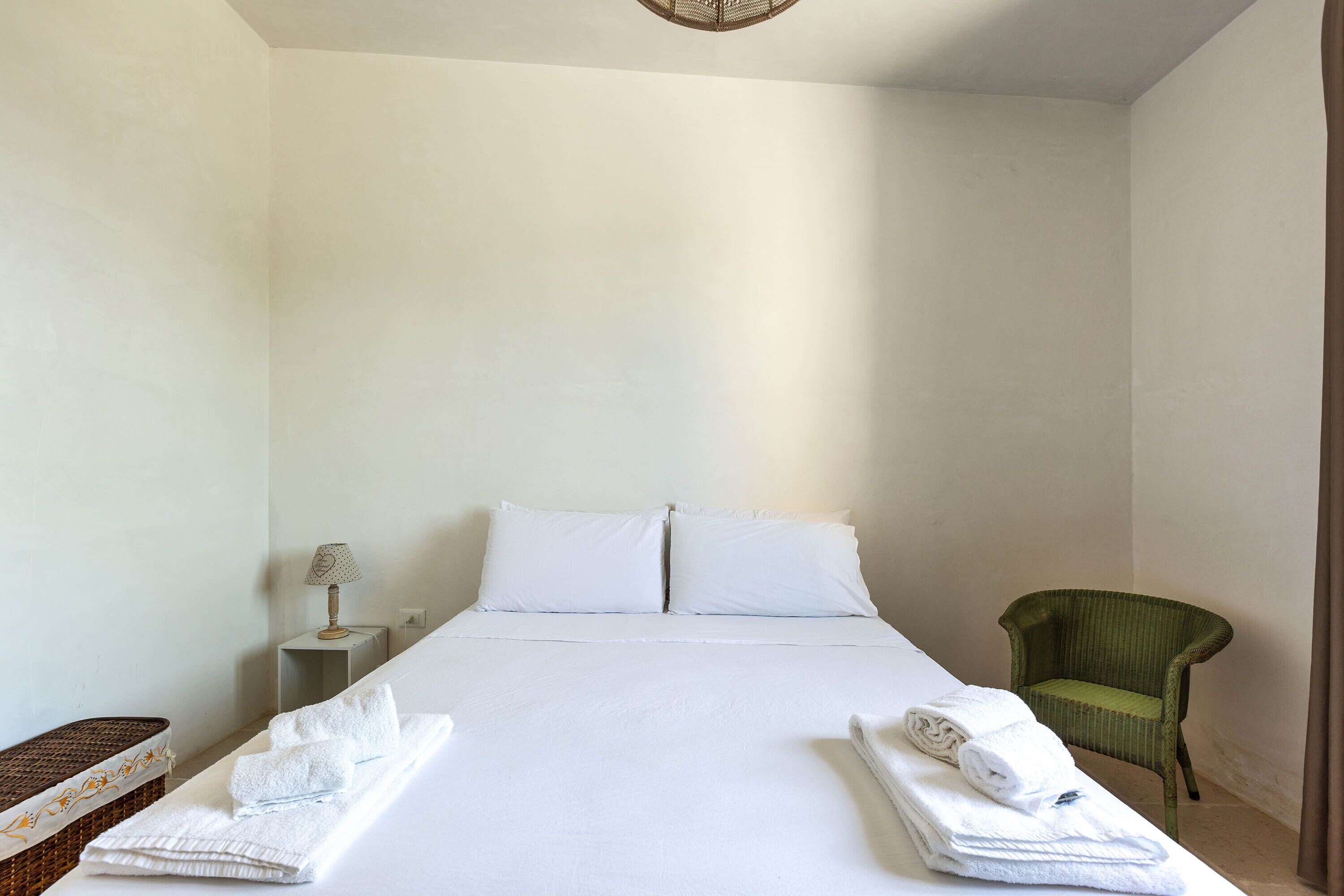 A neatly made bed with white linens and pillows, a small bedside table with a lamp, and a green chair.