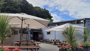 An outdoor seating area with picnic tables and umbrellas, surrounded by potted plants and a building with a sign. - Charming Two Bed Cottage Glan Gwna Holiday Park (Caernarfon)