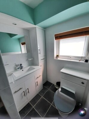 A compact bathroom with a white vanity, a toilet, and a window with blinds.