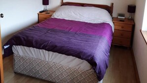 A bedroom with a bed, two bedside tables, a nightstand, a wall-mounted lamp, and a painting. - Charming Two Bed Cottage Glan Gwna Holiday Park (Caernarfon)