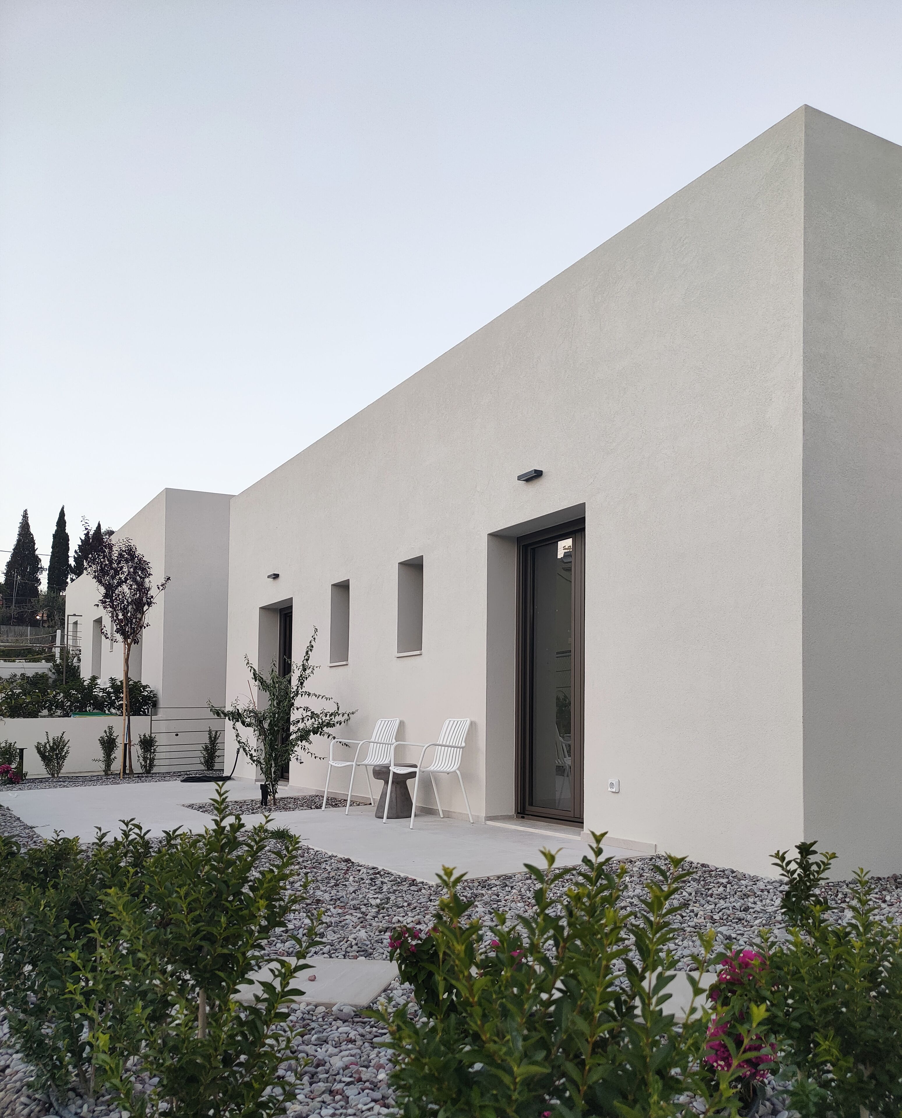 A modern building with a flat facade, a door, and a small patio area with chairs and plants.