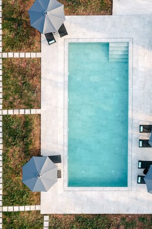 Et rektangulært pool med flisebelagt dæk hele vejen rundt. Der er to parasoller og to liggestole på terrassen.