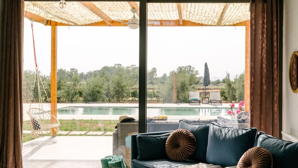 A living room with a sofa, a small round table, and a view of a swimming pool and greenery outside. - Irissa Suites (Nafplio)