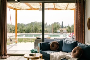 En stue med sofa, et rundt bord og udsigt til swimmingpool og grønne områder.