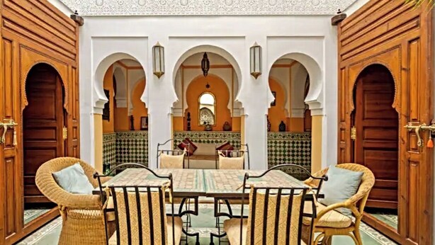 Ein traditionelles marokkanisches Interieur mit einem zentralen Tisch und Stühlen, Bögen und ornamentierten Fliesen.