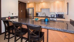 A modern kitchen with granite countertops, wooden cabinets, a stainless steel refrigerator, a coffee maker, a kettle, and two bar stools. - Three Kings 37 2 Bedroom Condo (Park City)