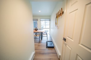 Un pasillo estrecho con suelo de madera, una puerta blanca a la derecha y un comedor al fondo con una mesa y sillas.