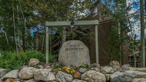A stone marker with "WILD IRISHMAN" inscribed, surrounded by rocks and flowers, with a wooden pergola overhead and buildings in the background. - Wild Irishman 1054 (Keystone)