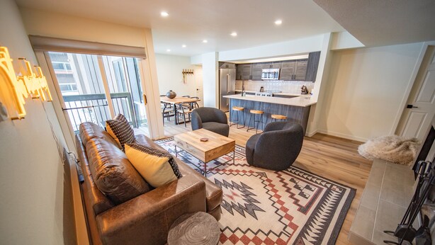 A modern living room with a sofa, chairs, and a coffee table. There is a kitchen area with a refrigerator, stove, and bar stools. - Wild Irishman 1054 (Keystone)