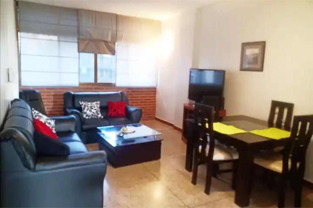 A living room with a sofa, coffee table, and a TV. There is a dining table with chairs and a window with blinds.