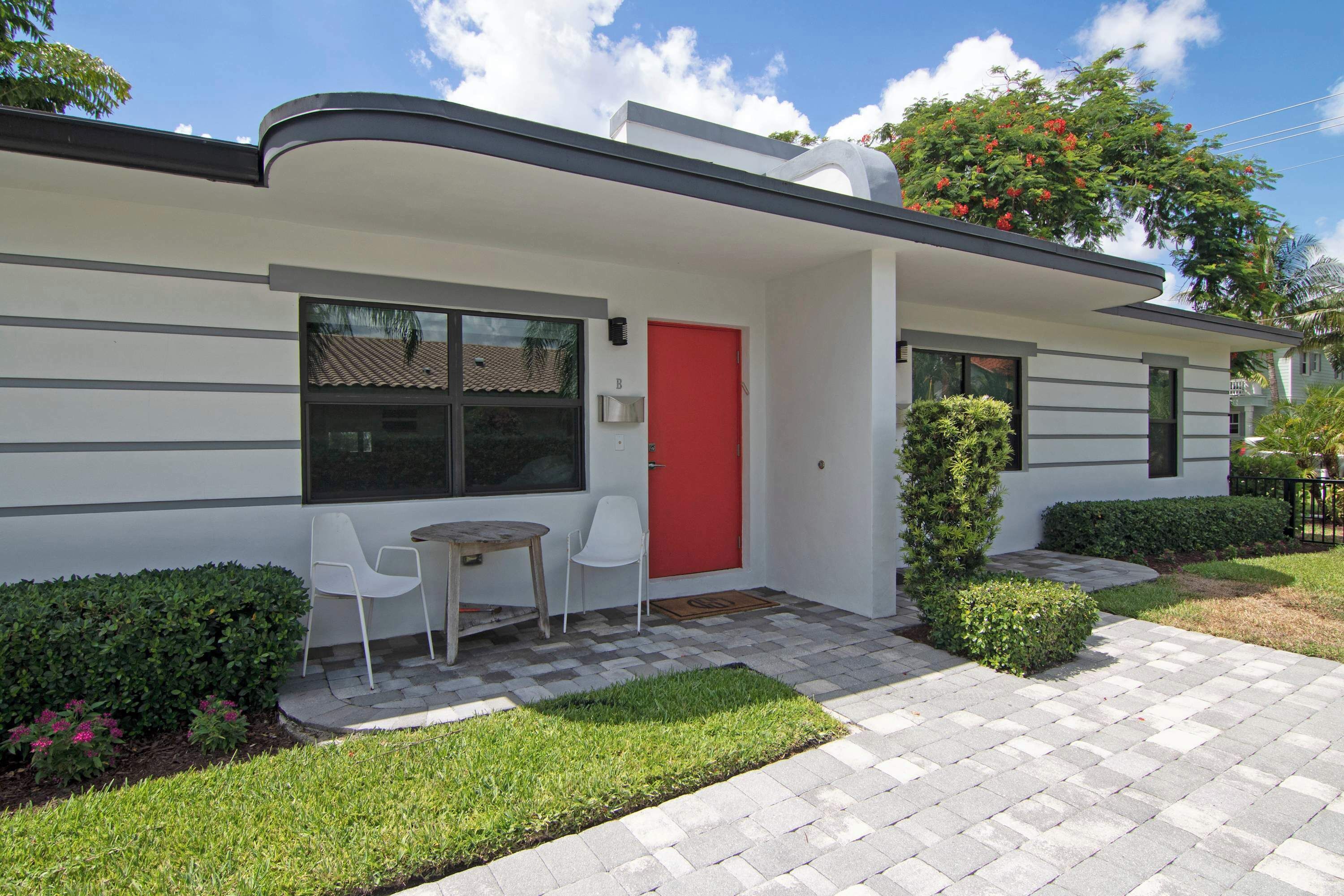 Une maison moderne avec une porte rouge, deux chaises blanches et une petite table à l’extérieur.
