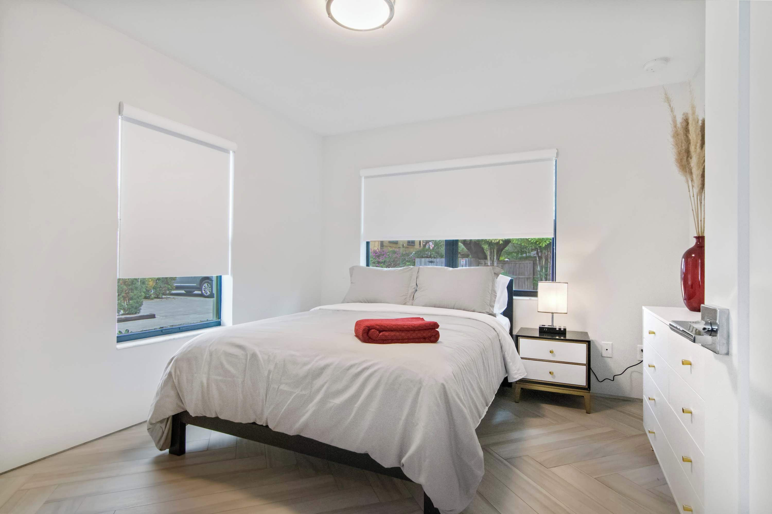 A bedroom with a bed, bedside table, lamp, dresser, and a window with a view.