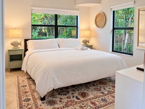 Un dormitorio con una cama grande, dos mesitas de noche, una lámpara auxiliar y una alfombra estampada, con ventanas que dan a zonas verdes.