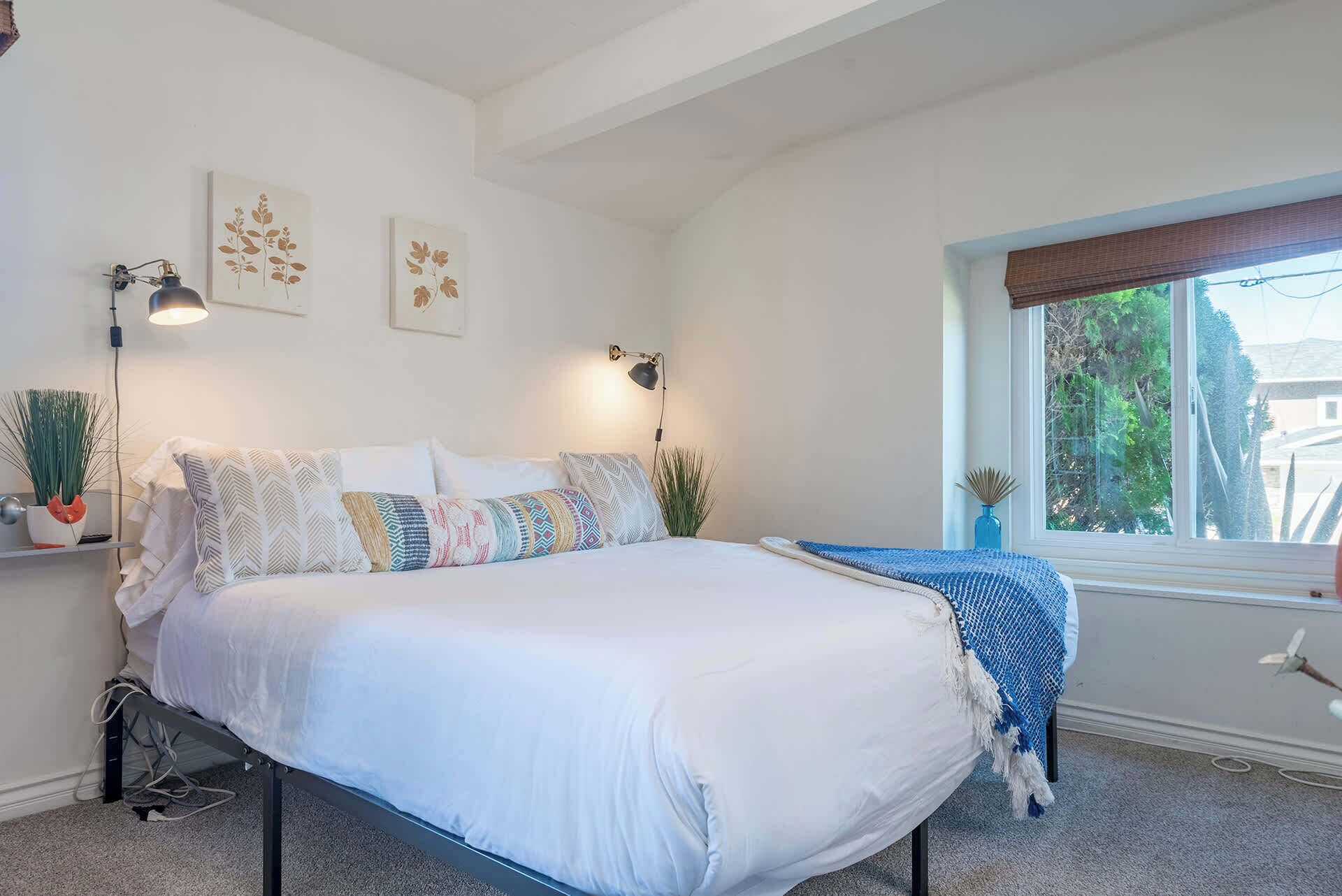 Un dormitorio con cama, dos lámparas de pared, una ventana con vistas y una planta en la mesita de noche.