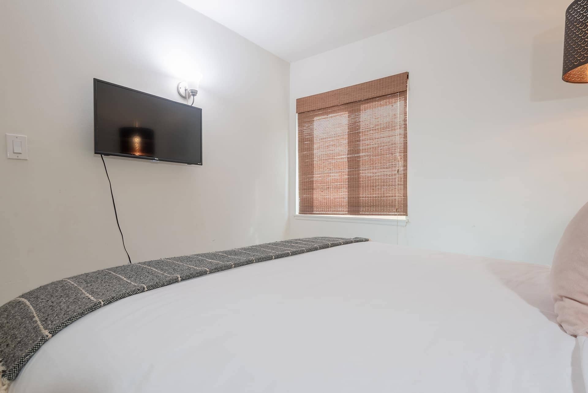 A bedroom with a bed, a mounted TV, a window with blinds, and a power outlet.