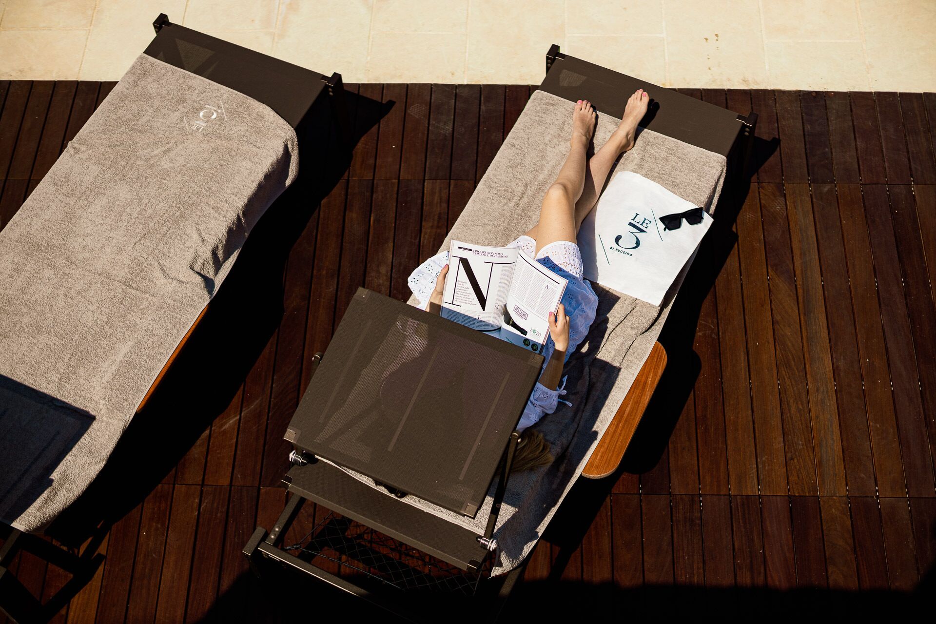 A person lying on a sun lounger with a magazine, another sun lounger beside it, and a wooden deck.