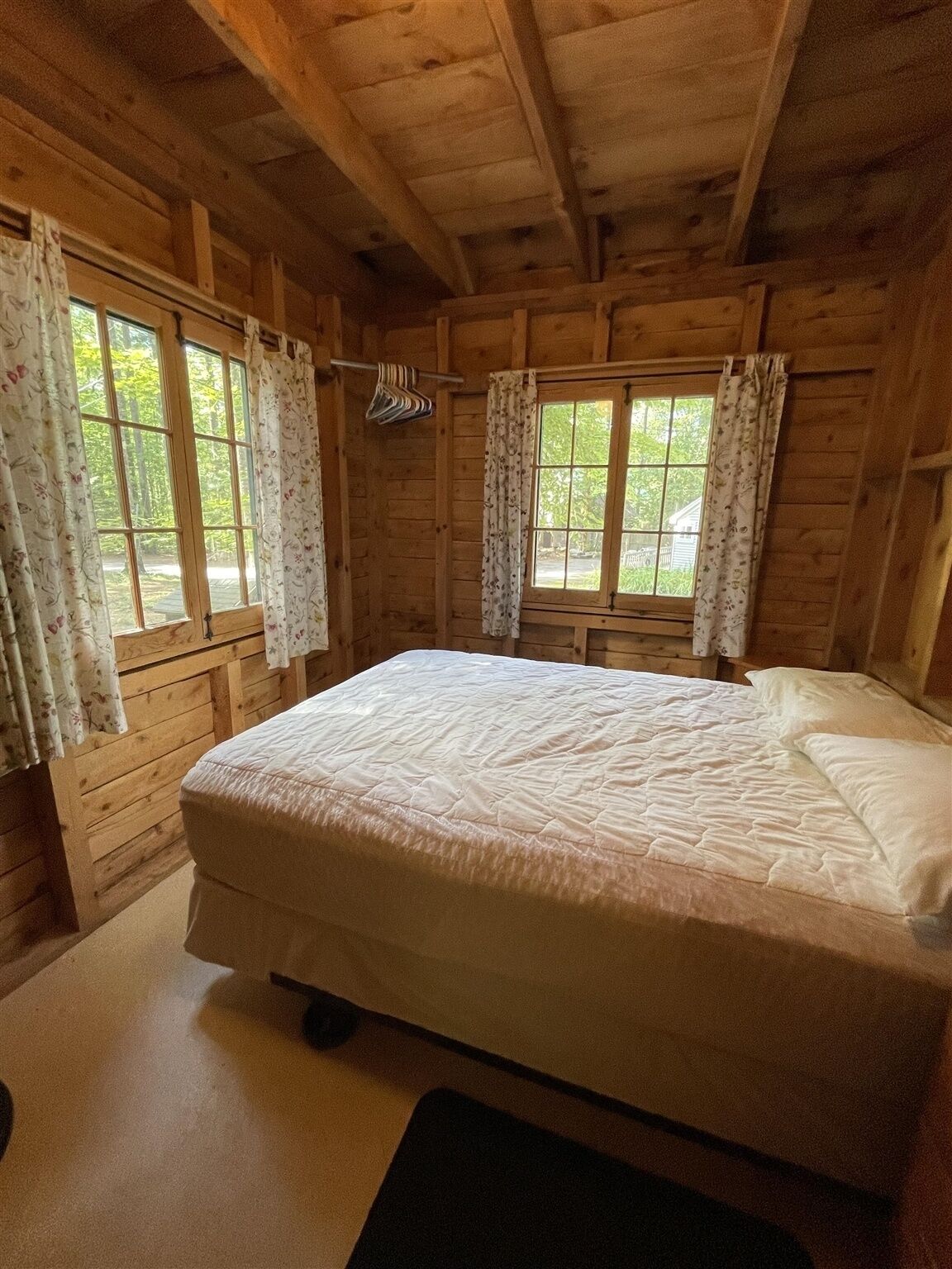 A wooden cabin bedroom with a large bed, two windows with curtains, and a wooden ceiling.