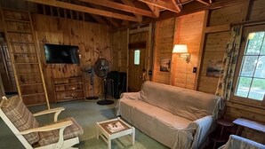 A cozy log cabin interior with a sofa, armchair, and ottoman, a television, and a fan.