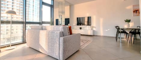 A modern living room with a grey sofa, a dining area with a table and chairs, a TV mounted on the wall, and large windows offering a city view.