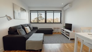 A modern living room with a black sofa, a white table, a grey rug, a TV, and a large window with a view of trees and buildings. - Downtown Stylish Apartment Garage by Vacationy (Porto)