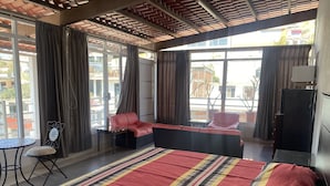 Habitación de hotel con una cama grande, un sofá, una silla y una mesita. Cuenta con ventanas amplias con cortinas, y vistas al exterior.