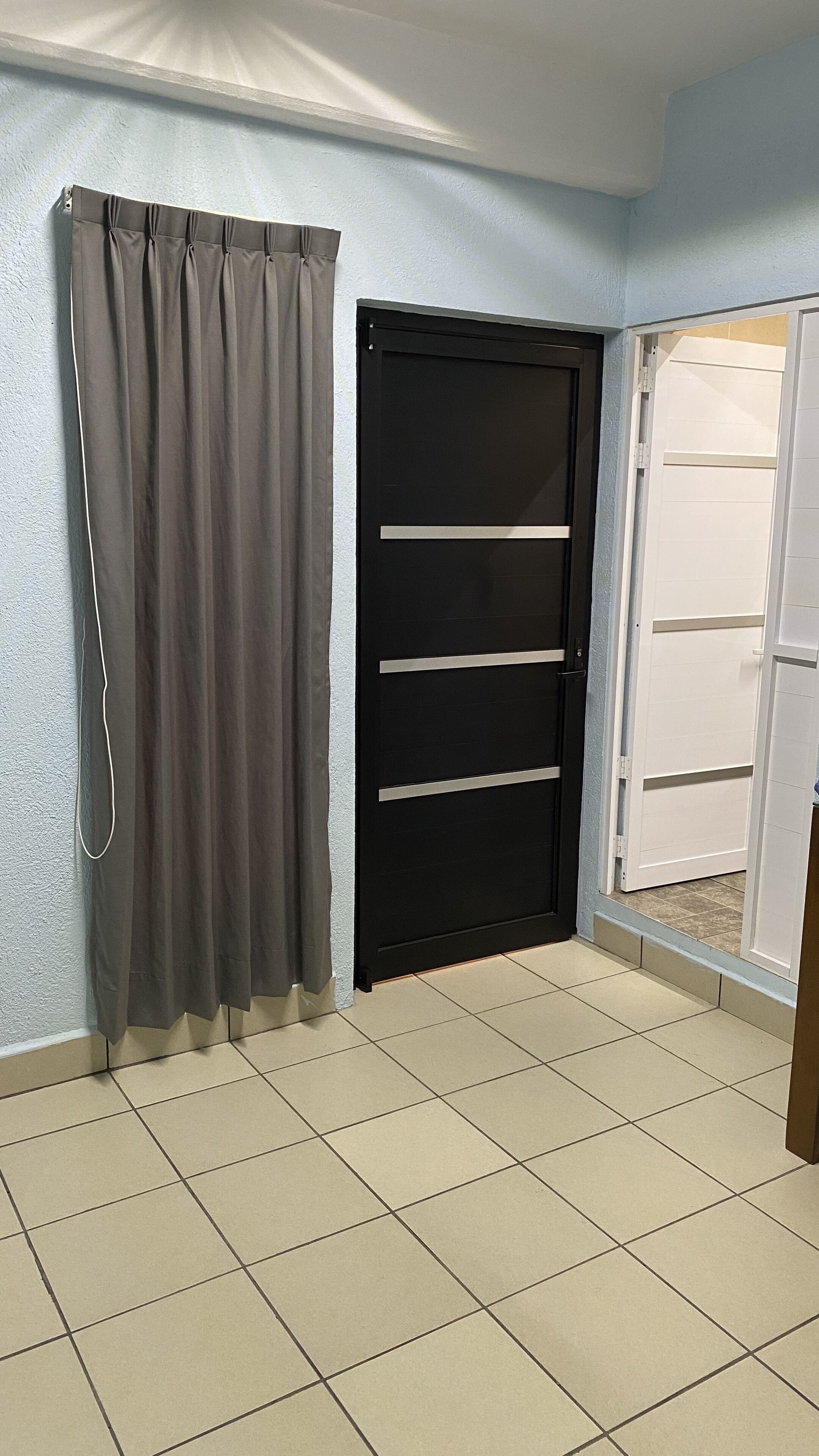 Une pièce avec une porte noire fermée, un rideau gris et un sol carrelé.