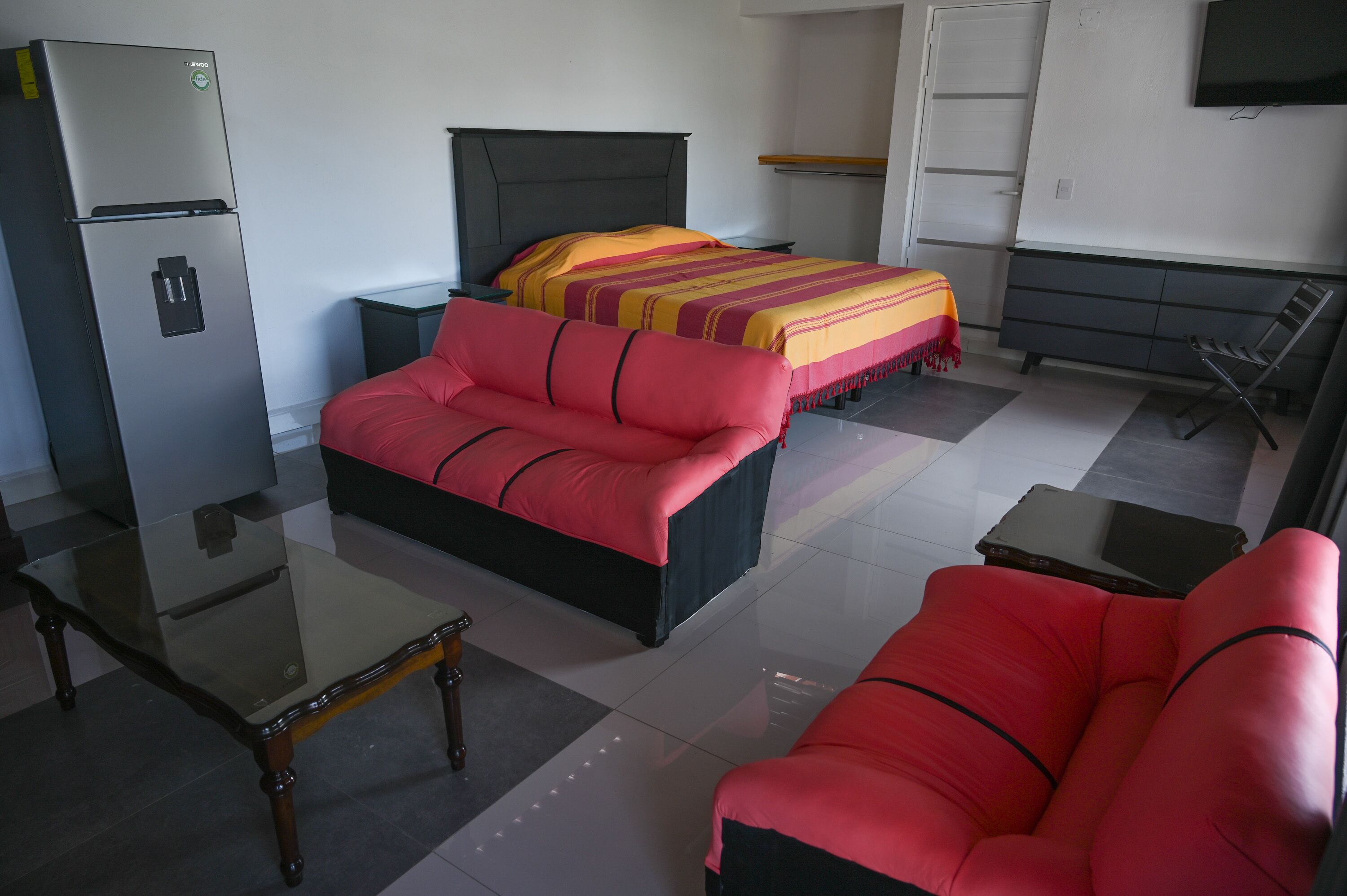 A hotel room with a bed, a red sofa, a glass coffee table, a refrigerator, a TV, and a wardrobe.