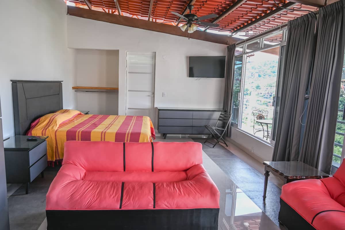 A bedroom with a bed, a red sofa, a desk, a TV, and a window with curtains.