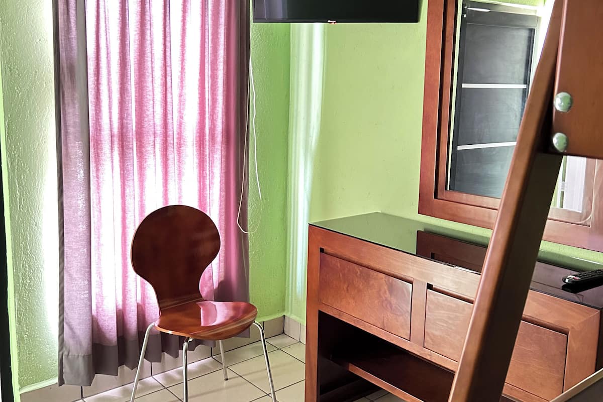 A room with a green wall, a wooden desk, a chair, a television mounted on the wall, and a window with curtains.