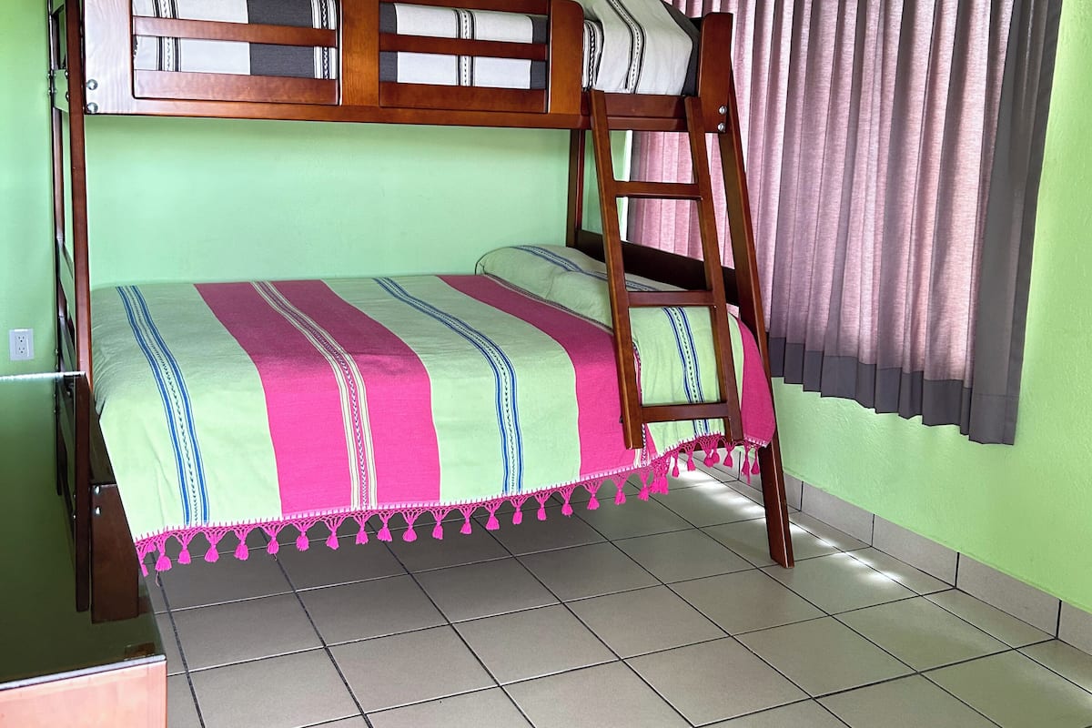 A bunk bed with a wooden frame and a colorful bedspread, a window with curtains, and a tiled floor.