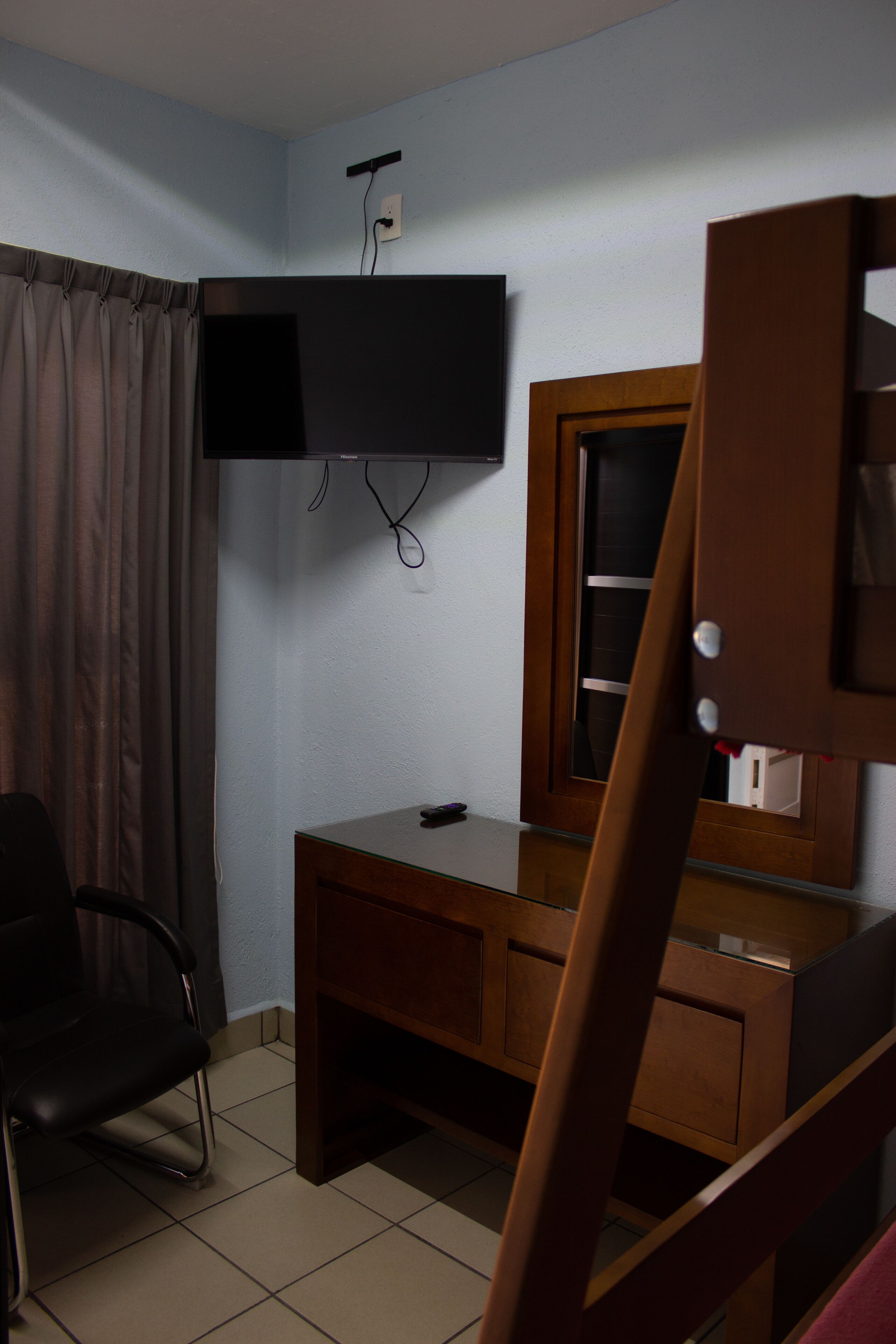 A room with a wooden desk, a chair, a television mounted on the wall, and a window with curtains.