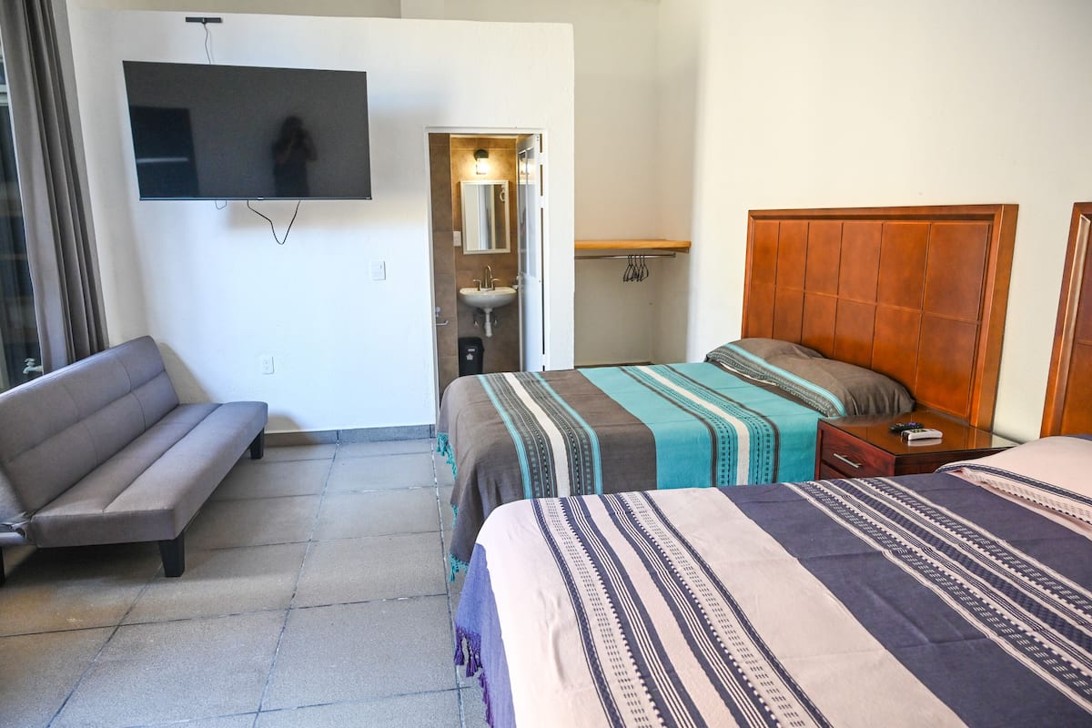 A hotel room with a sofa, a bed with a wooden headboard, a TV mounted on the wall, and a bathroom visible through an open door.