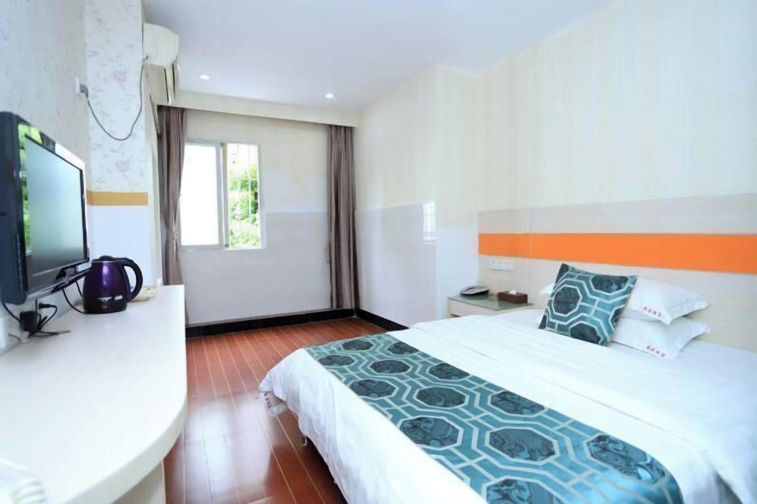 A hotel room with a bed, a desk with a television and a coffee maker, a window with curtains, and a nightstand with a lamp.