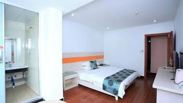A hotel room with a bed, a desk, a chair, a glass shower enclosure, and a small bathroom.