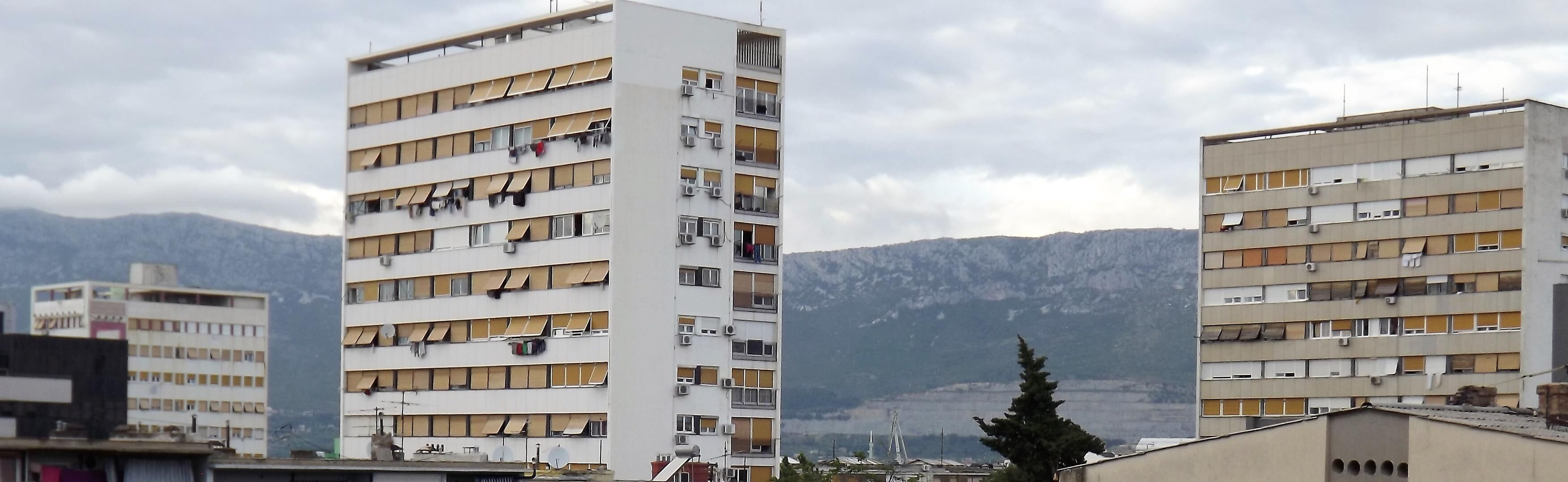 A multi-story building with balconies, a mountainous backdrop, and a clear sky.