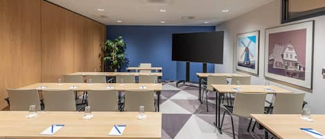 A conference room with multiple tables, chairs, a large screen, and a potted plant.