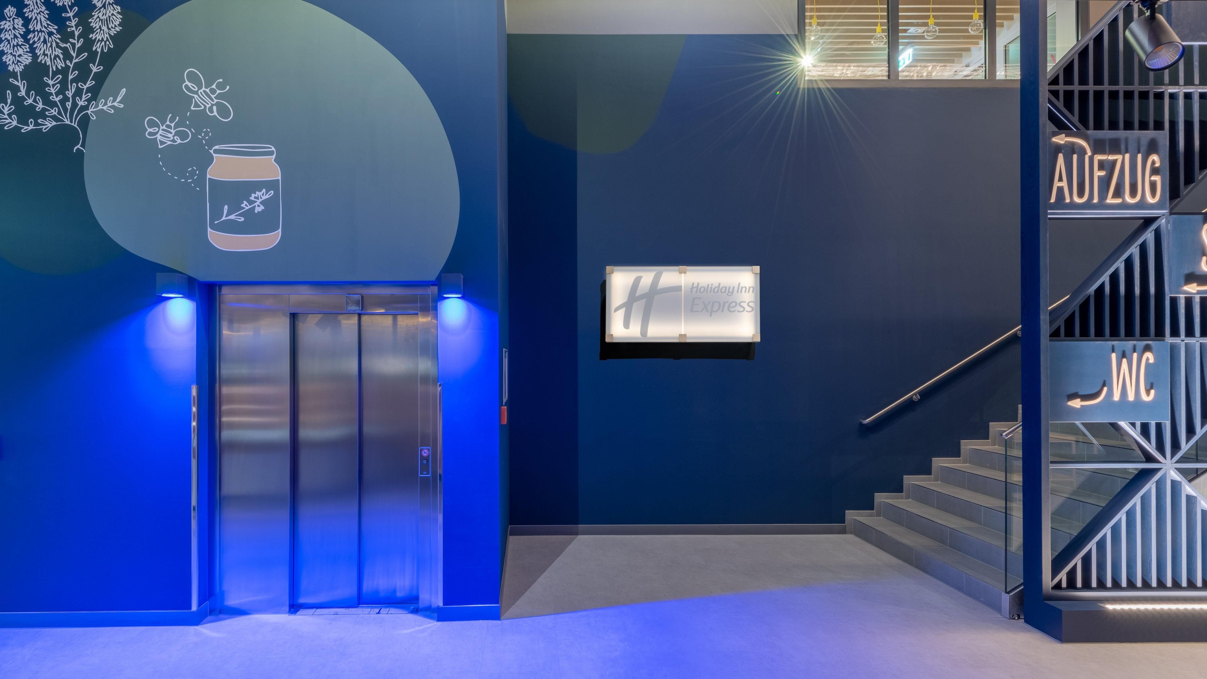 En moderne hotellobby med en blå væg, elevator, trappe og skilt der viser vej til toiletterne.