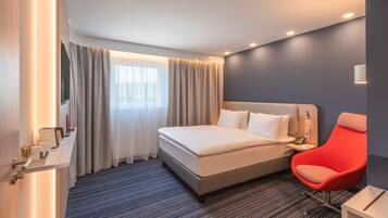 Une chambre d’hôtel moderne avec un grand lit, un fauteuil rouge, une table de chevet et une fenêtre avec des rideaux.