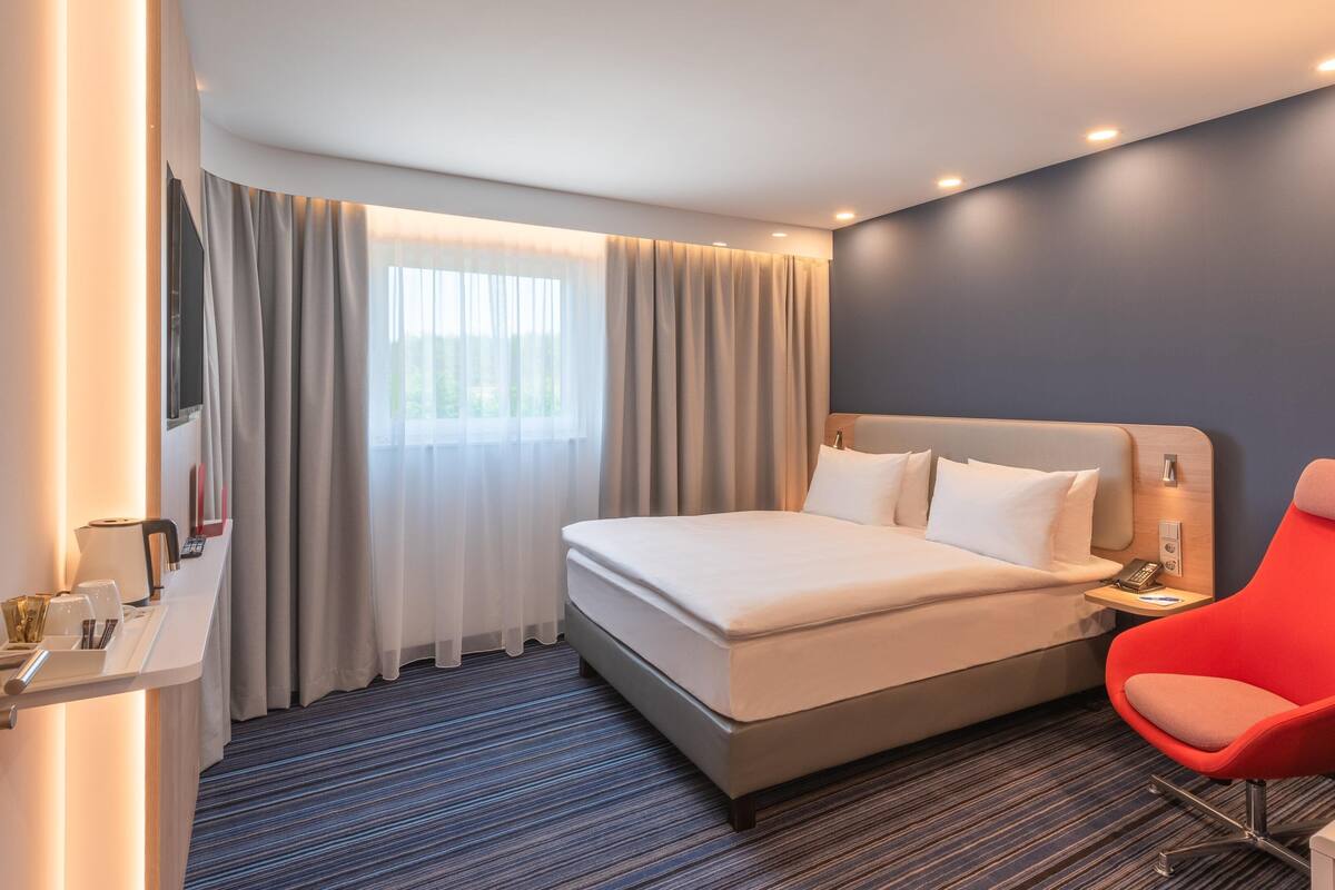 Une chambre d’hôtel moderne avec un grand lit, un fauteuil rouge, une table de chevet et une fenêtre avec des rideaux.