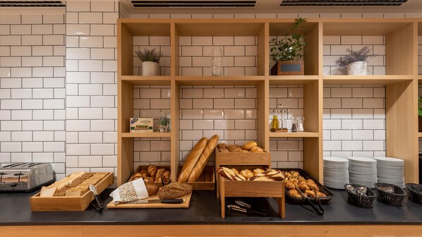 A bakery display with various breads and pastries, a toaster, and neatly stacked plates and baskets. - Holiday Inn Express Hamburg South A7 As42 (Egestorf)