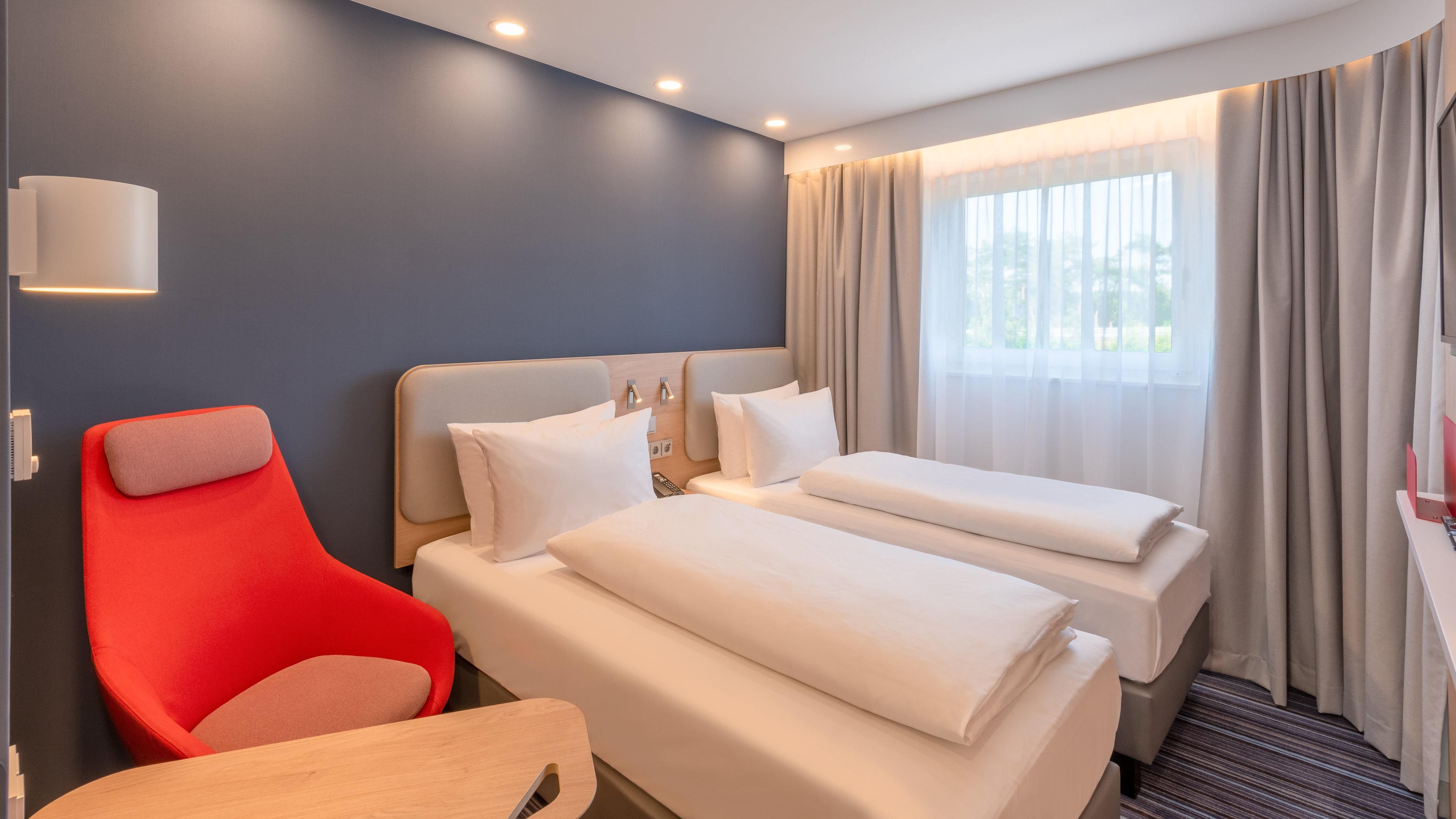 A hotel room with two beds, a red chair, a bedside table, and a window with curtains.