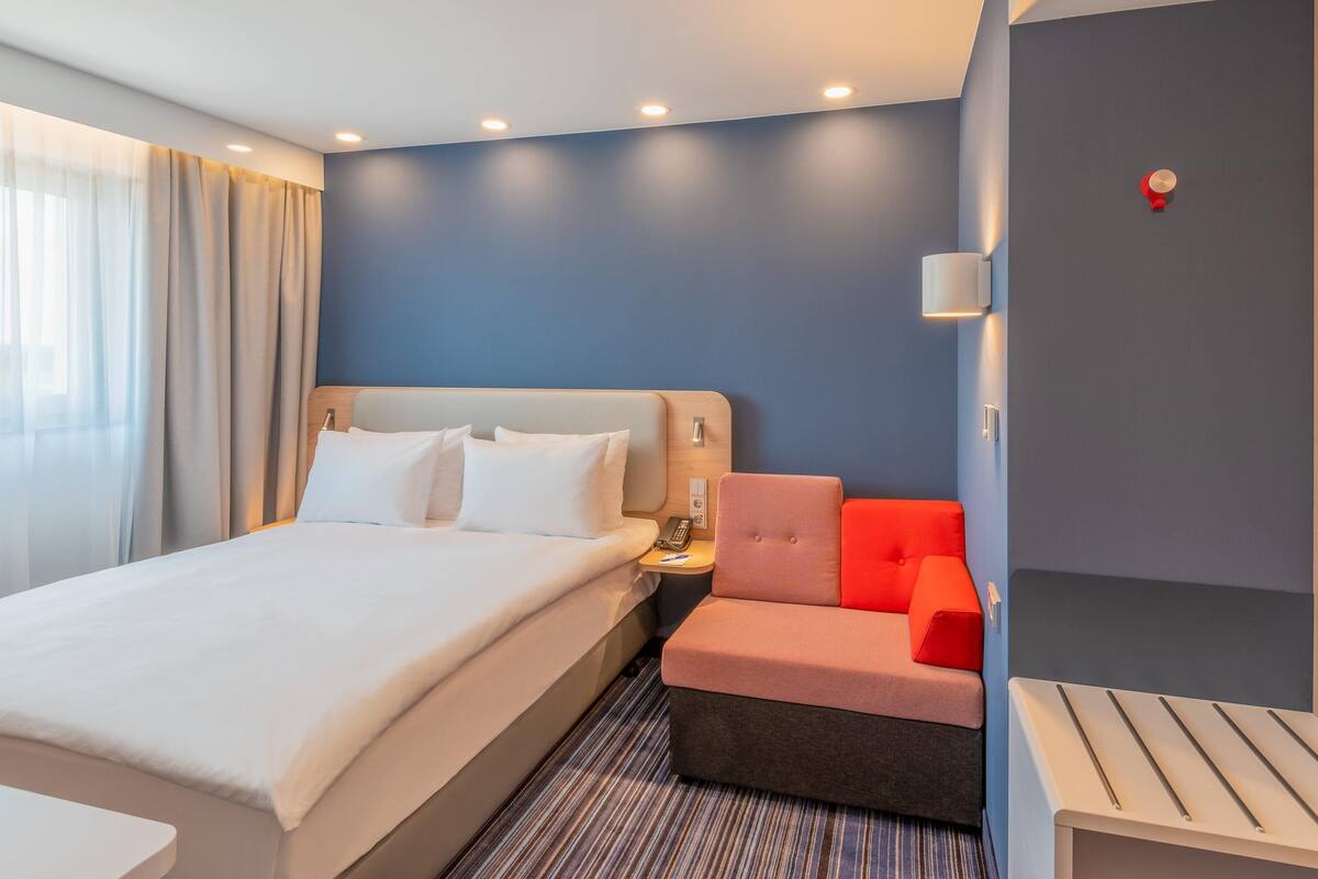 Une chambre d’hôtel moderne avec un grand lit, un bureau et un coin salon agrémenté d’un fauteuil rouge.