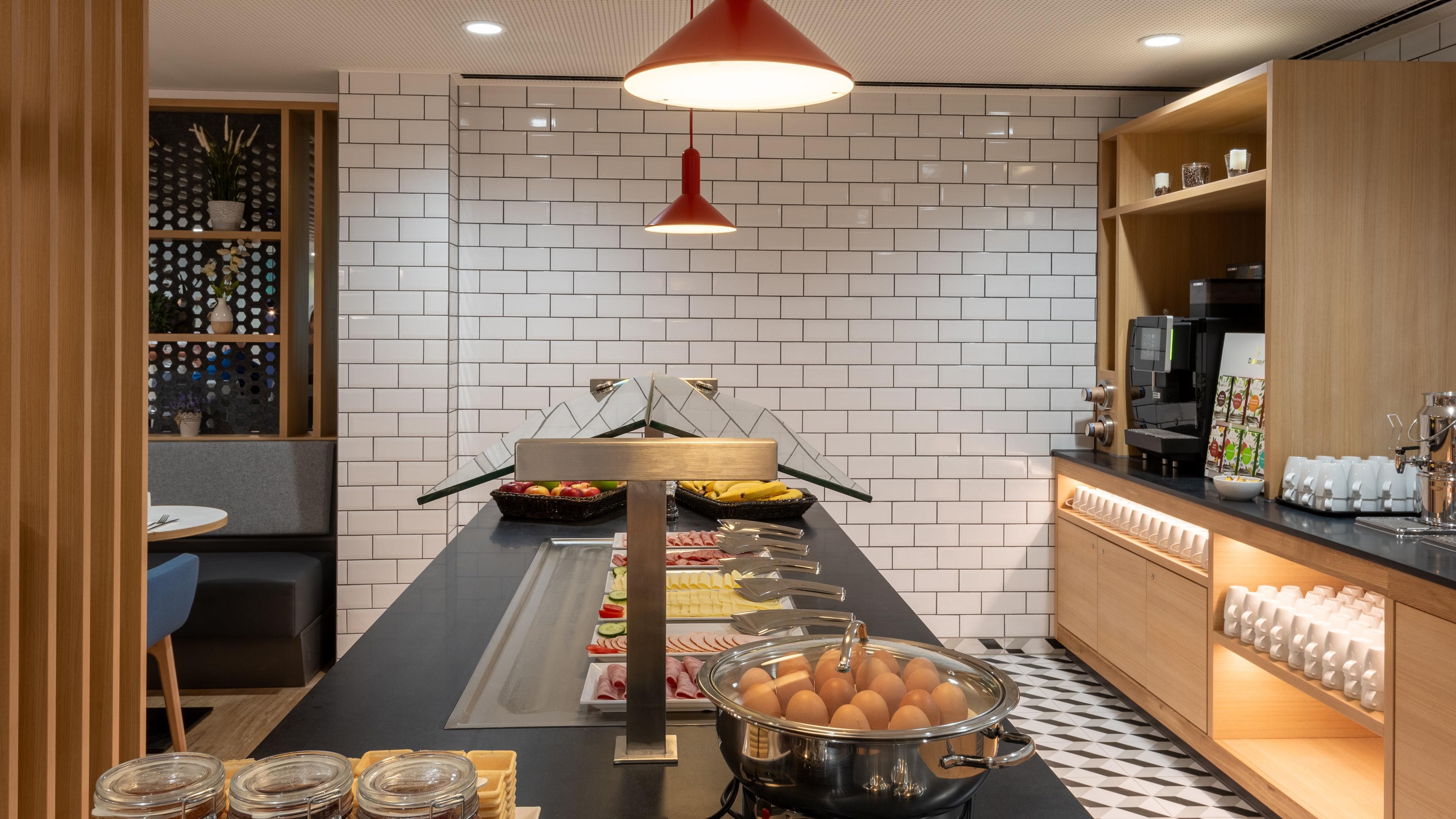 Montaje de bufé con diversos alimentos, una máquina de café y un área de estar con mesa y sillas.