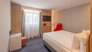 A modern hotel room with a large bed, a flat-screen TV mounted on the wall, a red chair, and a small table with a red cushion. - Holiday Inn Express Hamburg South A7 As42 (Egestorf)