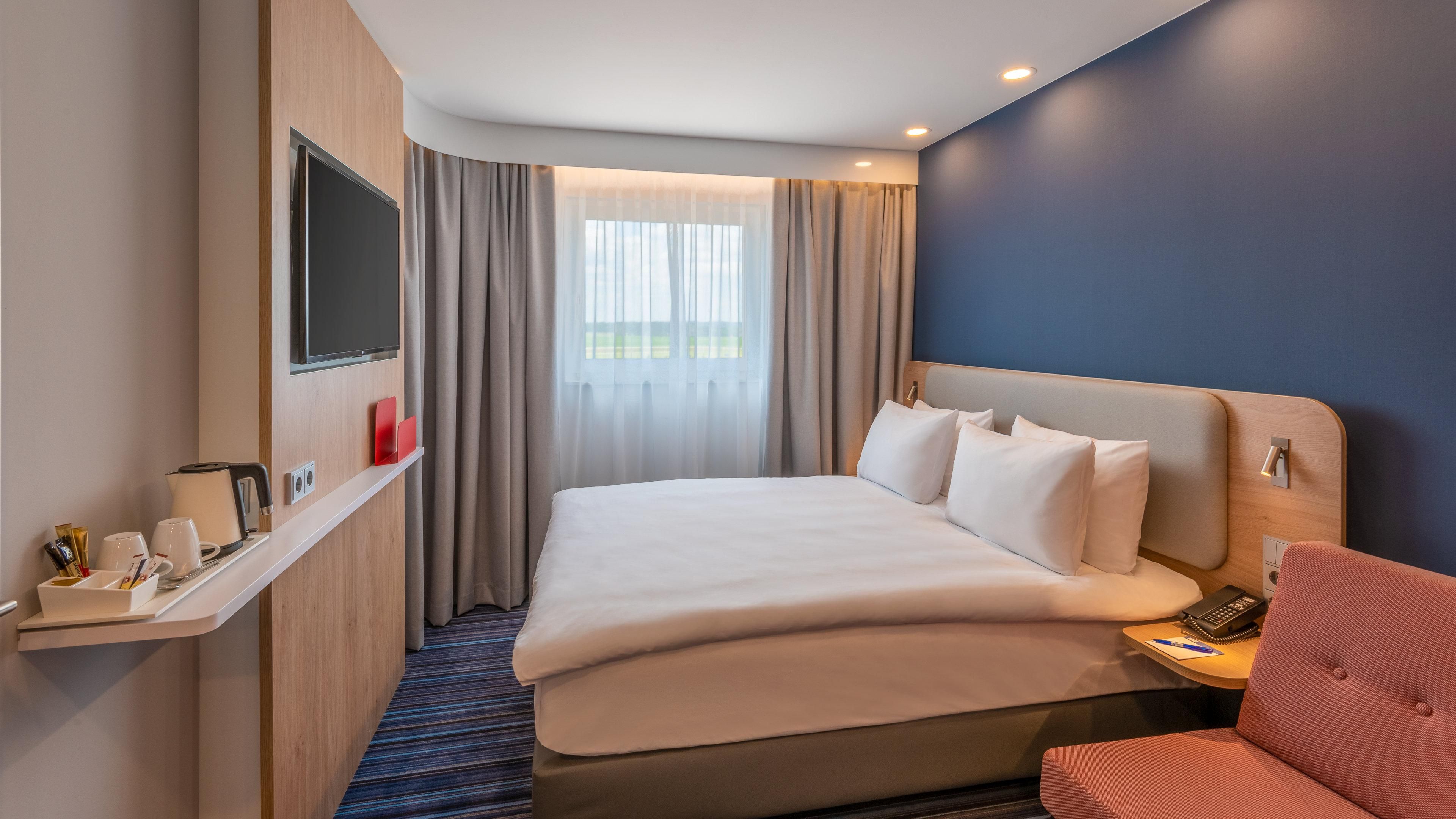 Una habitación de hotel con una cama grande, un televisor montado en la pared, una mesita de noche con un teléfono y una lámpara, y una ventana con cortinas.